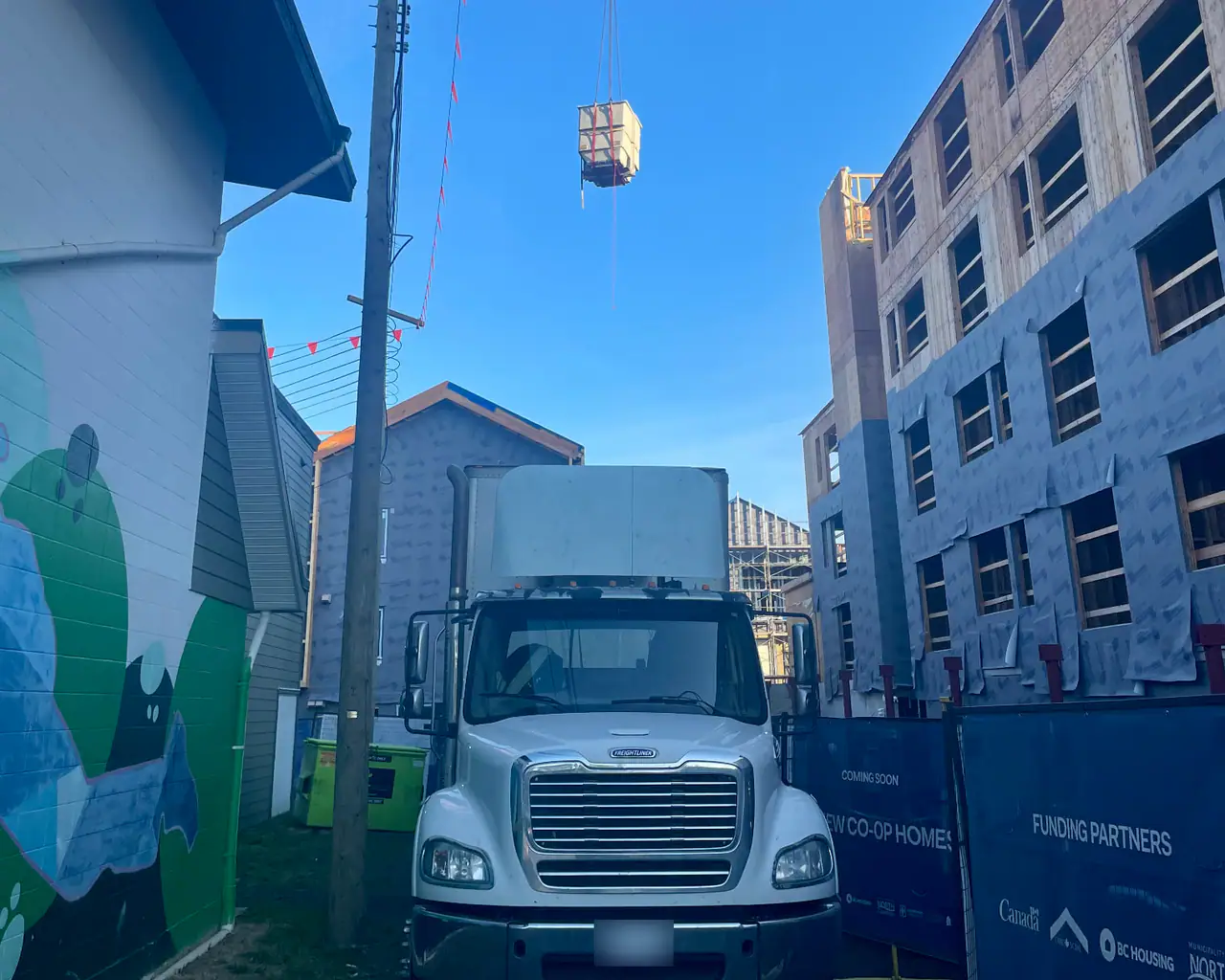 A white truck parked in a narrow lane between buildings under construction while a red tower crane hoists a load overhead