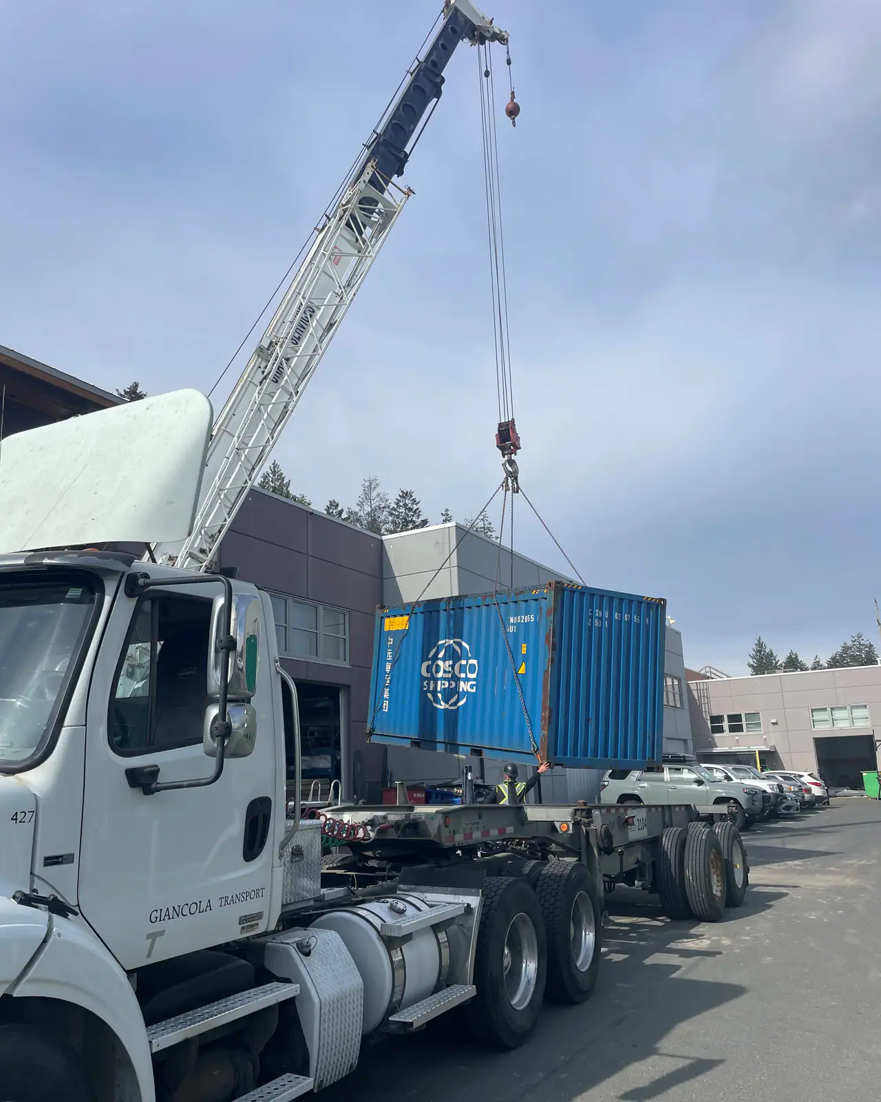 A mobile crane lifts a blue shipping container from a chassis while a worker in a hard hat steadies it, beside a white tractor