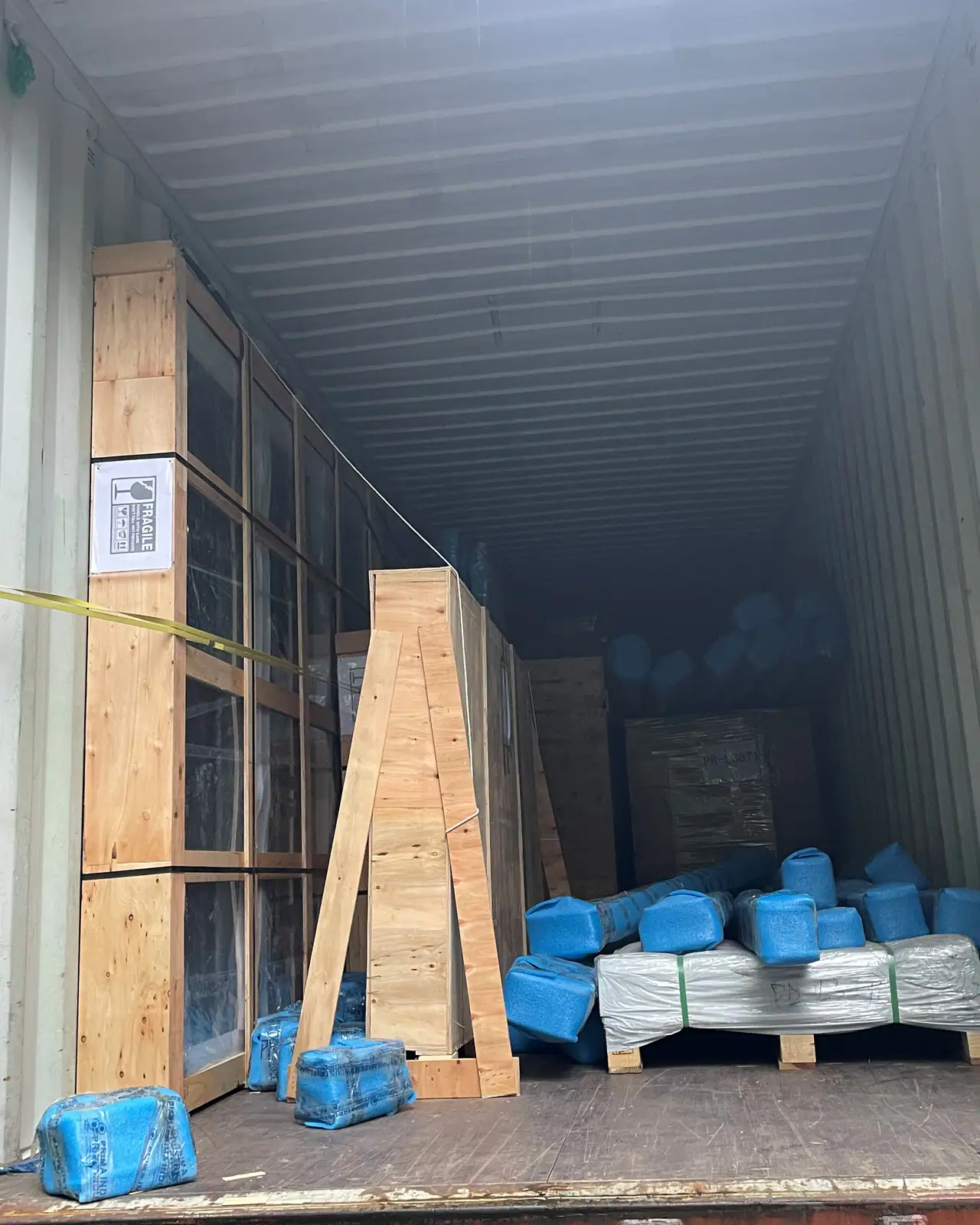 Tall timber-framed crates braced upright against the wall of a shipping container, with bundles wrapped in blue plastic on the floor beside them