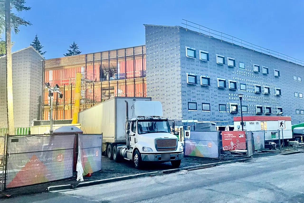 A white tractor and trailer parked on a building site in blue pre-dawn light, the unfinished building lit from inside