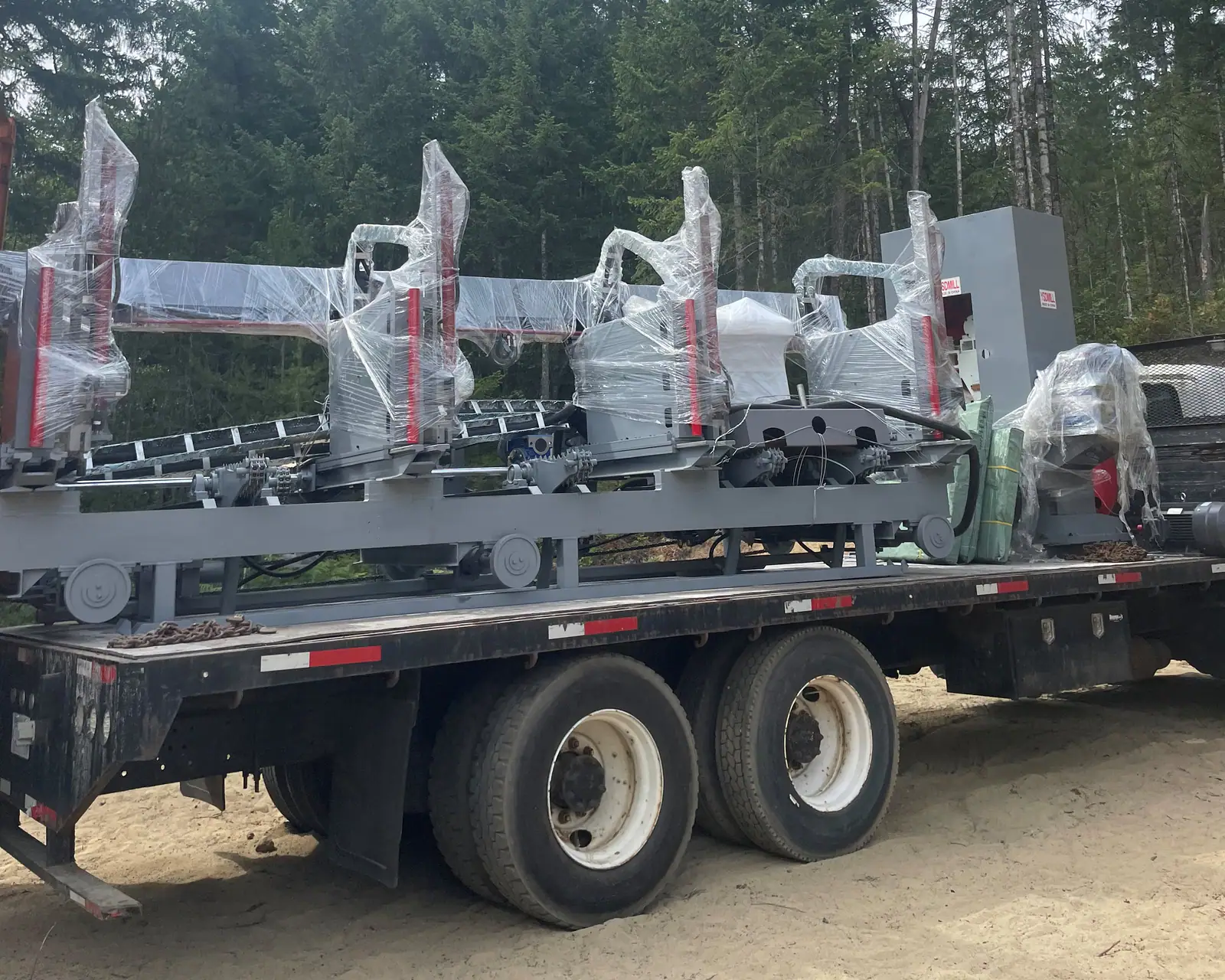Stretch-wrapped industrial machinery chained to the deck of a flat-deck truck on a sand lot among firs