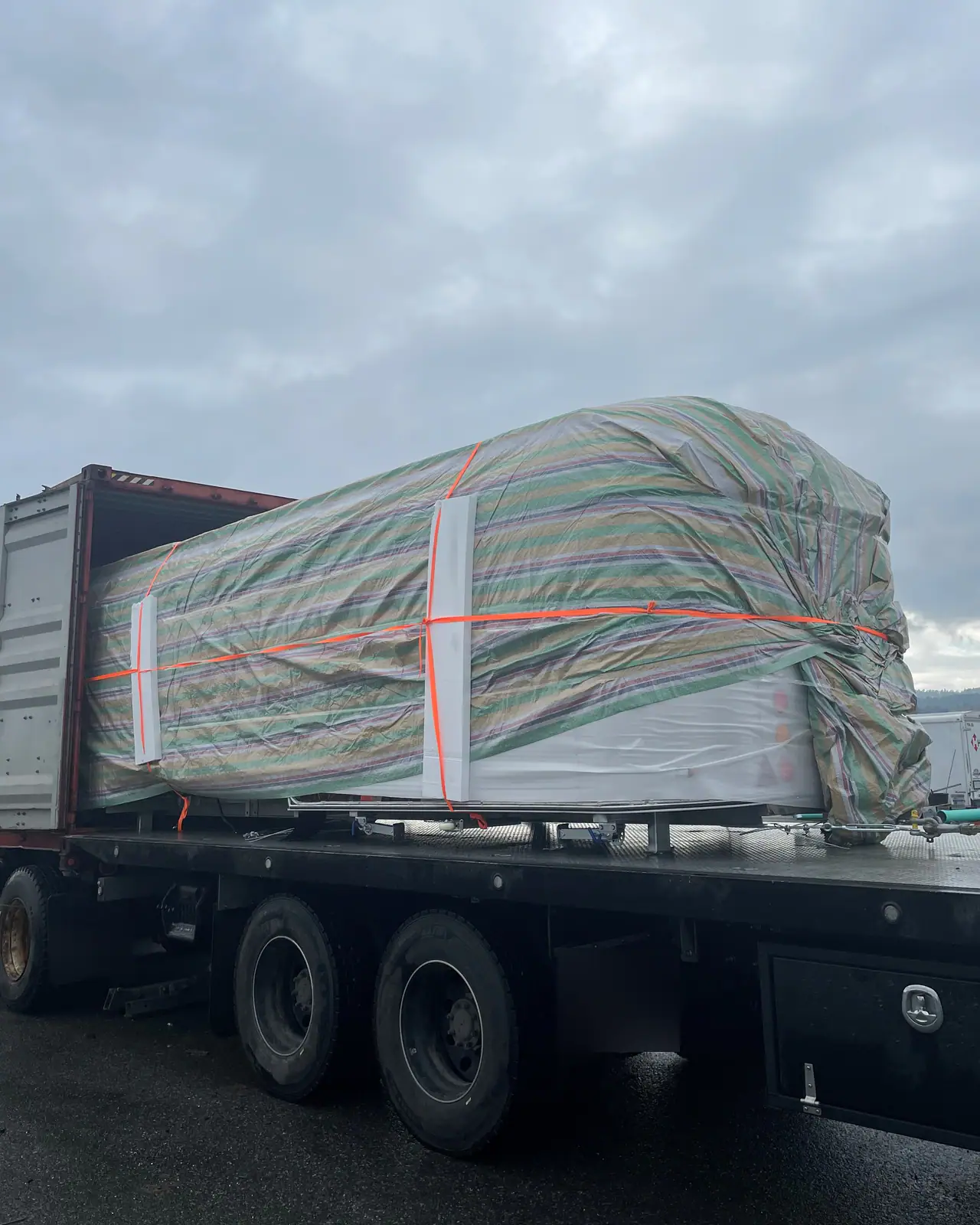 A large load wrapped in striped tarpaulin and orange strapping slides from an open shipping container onto the deck of a flat-deck truck under a heavy grey sky