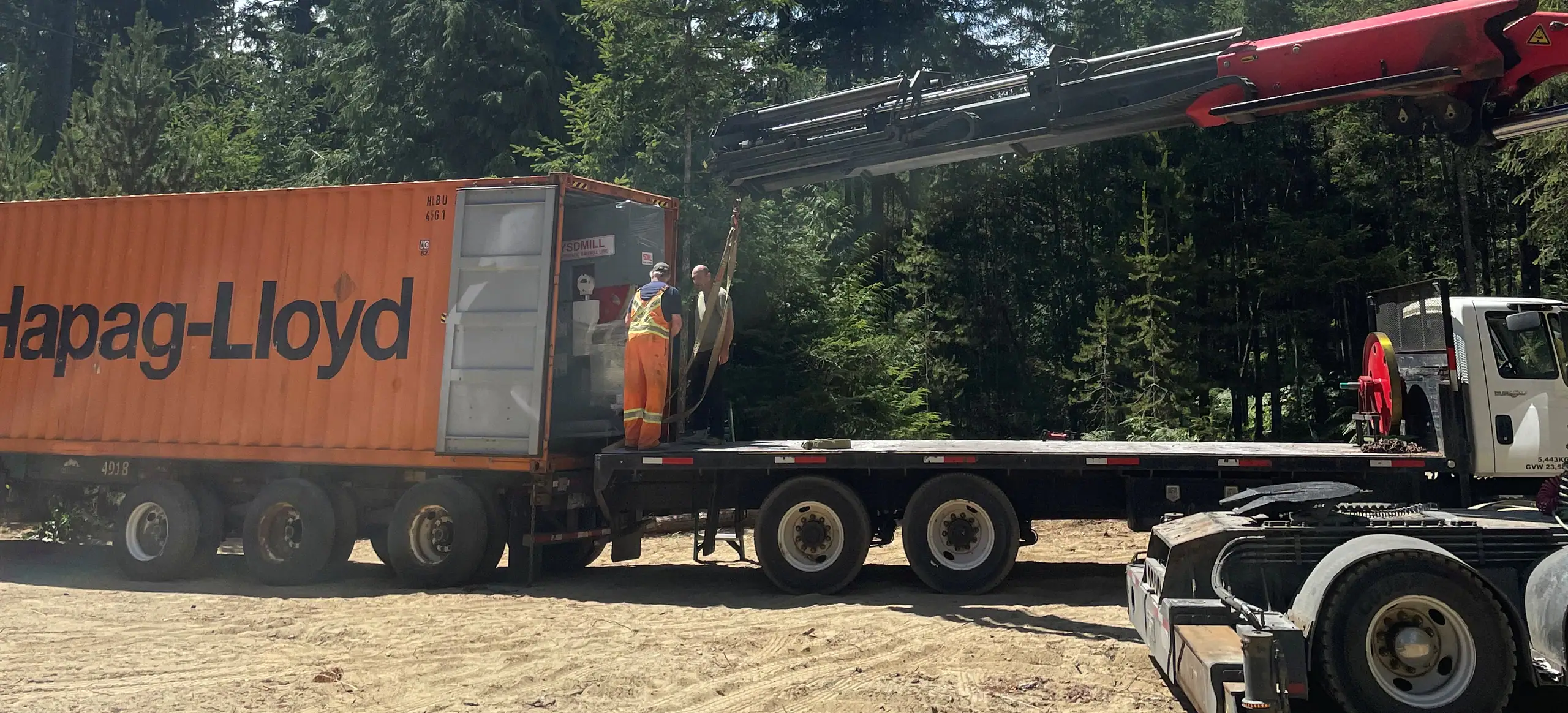 Two workers at the open doors of an orange shipping container on a chassis while a crane truck reaches over it, on a sand lot ringed by tall firs