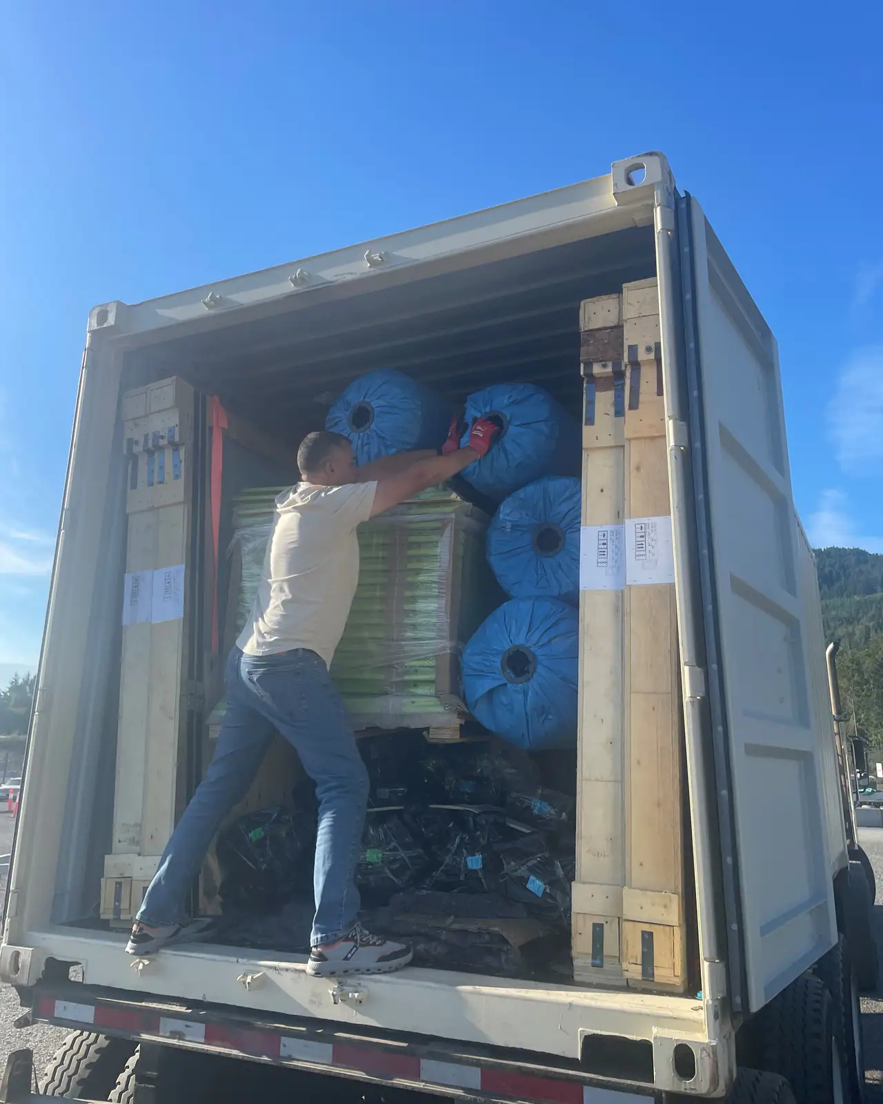 A man in a T-shirt and work gloves leans into a stack of wrapped rolls inside an open shipping container on a chassis