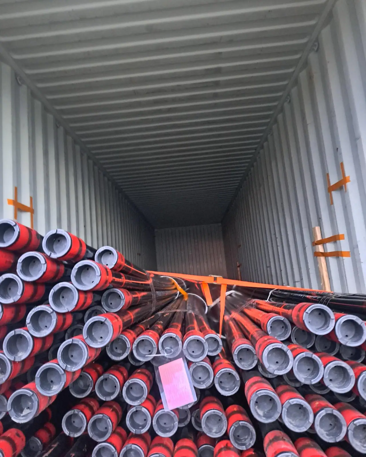 Red-painted steel pipe with grey end caps stacked in an open shipping container, braced with orange lashing