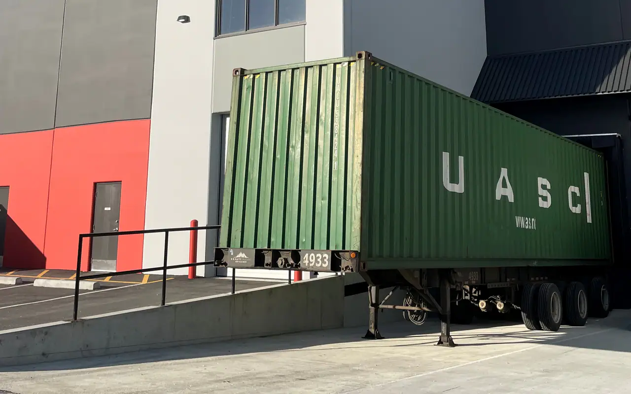 A green shipping container on a chassis backed up to a loading dock at a grey and white industrial building under a deep blue sky