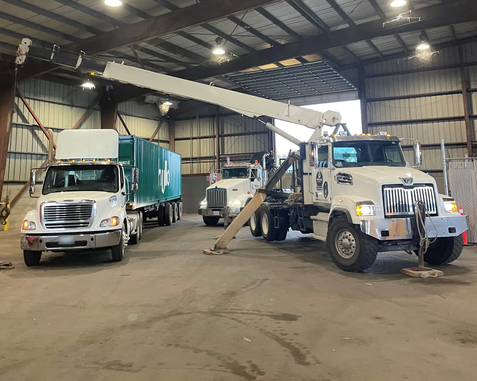 Two tractors and a boom truck under the roof of a large steel warehouse, one tractor coupled to a green container