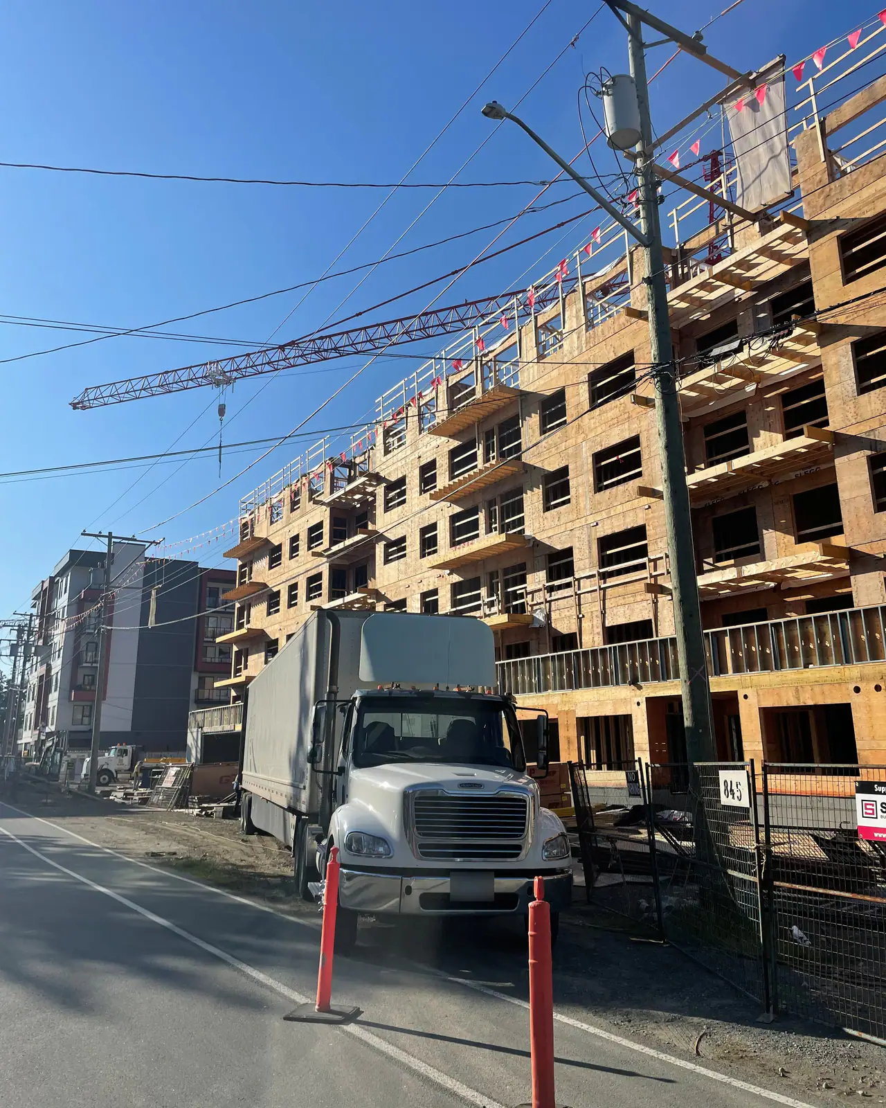 A white tractor and van trailer parked at the kerb beside a timber-frame apartment building under construction, a tower crane overhead