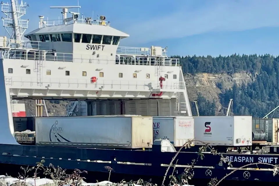 Drop trailers lined up on the open deck of a commercial barge, seen from a rocky shore with a forested bluff behind
