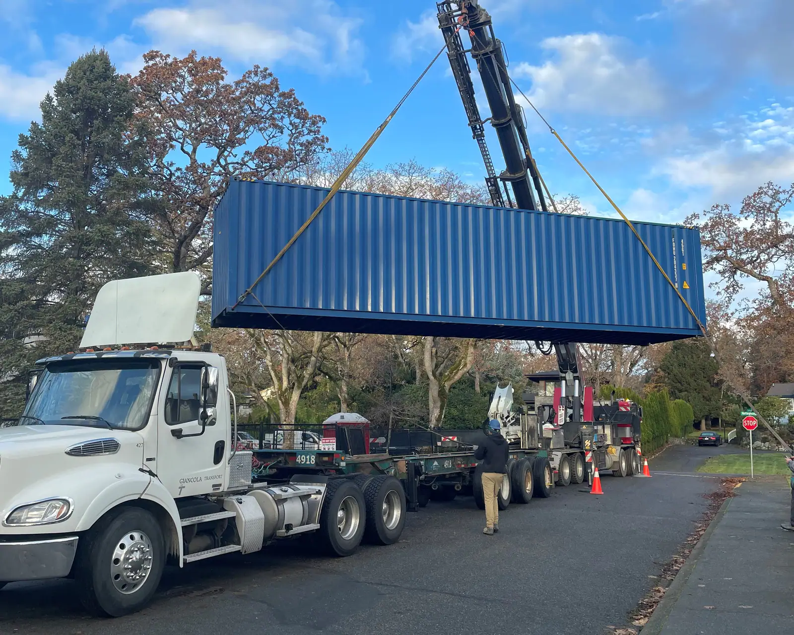 A blue shipping container lifted off a chassis by a crane truck on a quiet residential street, with a worker guiding it