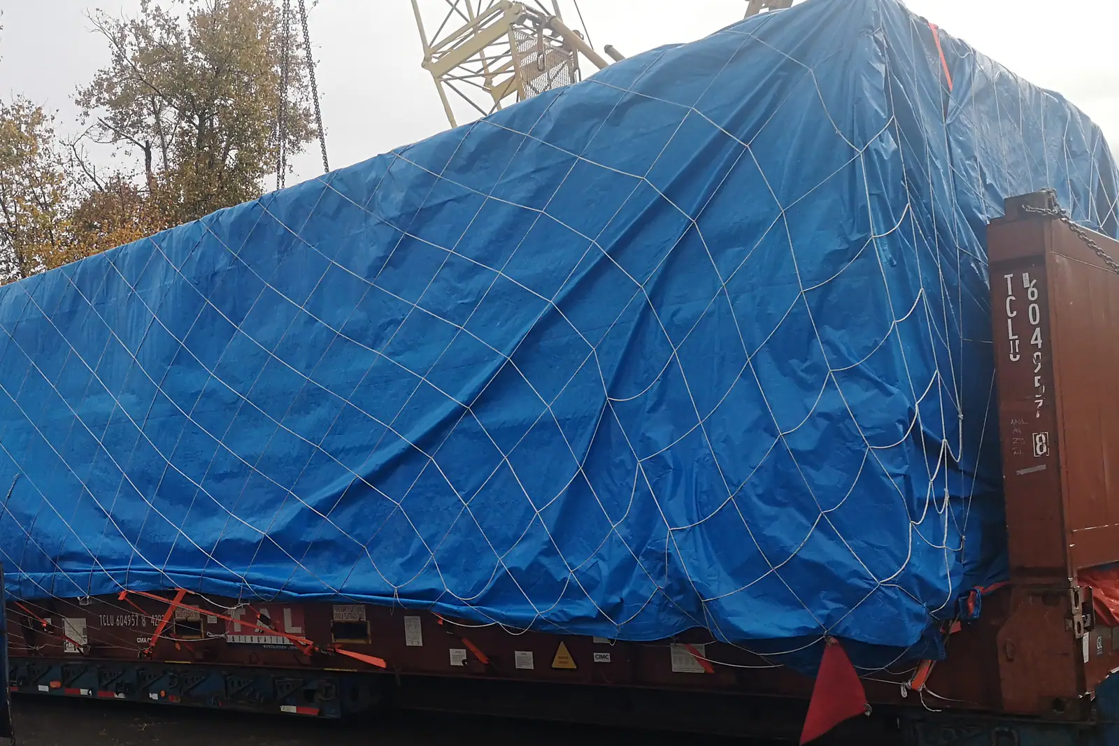 A tall load under a blue tarpaulin and cargo net on a flat-rack container, a red flag marking its overhang, in a yard beneath a crane