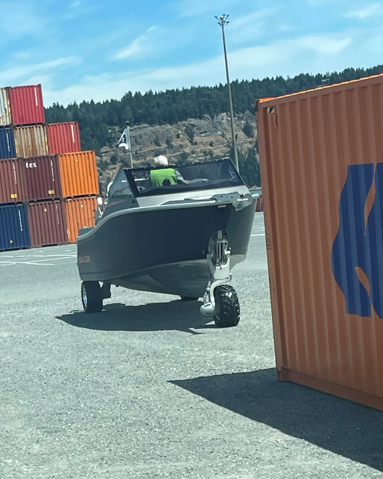 A grey amphibious boat drives on its own wheels across a container terminal, past a stack of shipping containers, with a forested bluff beyond