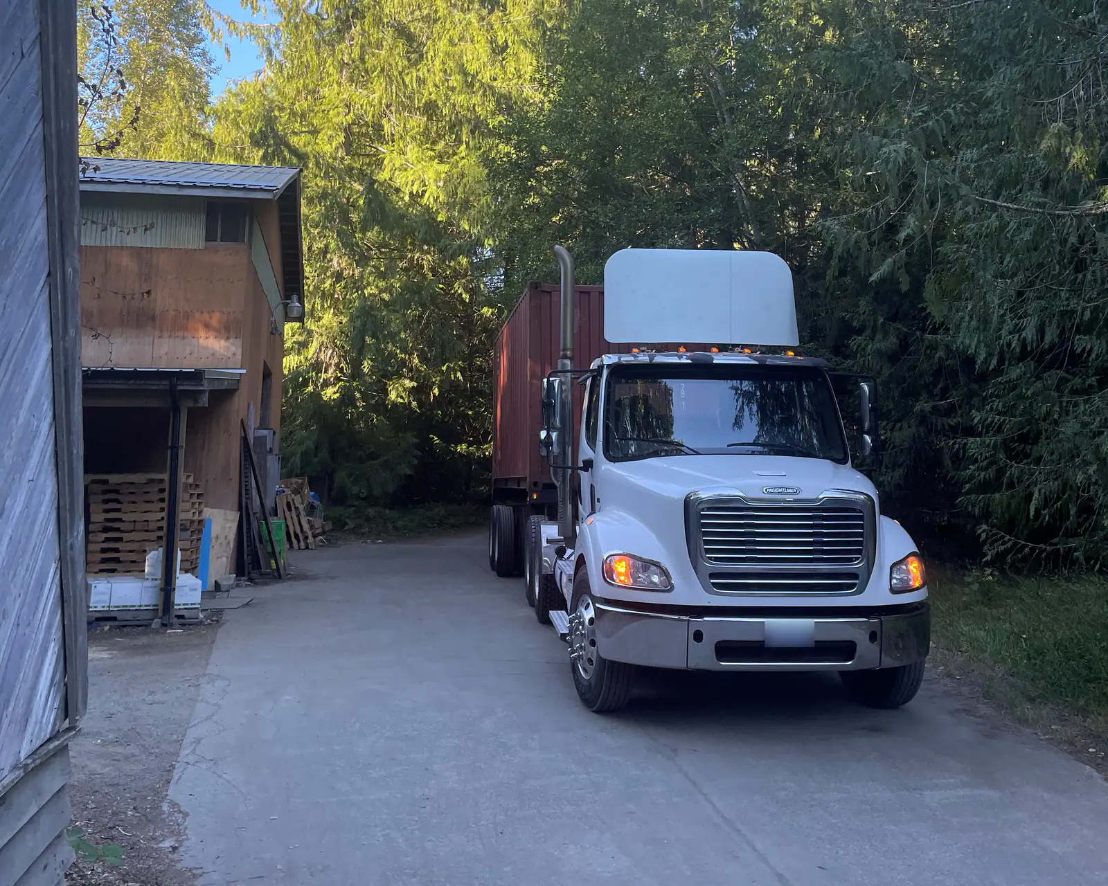 A day-cab tractor pulling a red container up a narrow driveway between a timber building and tall firs