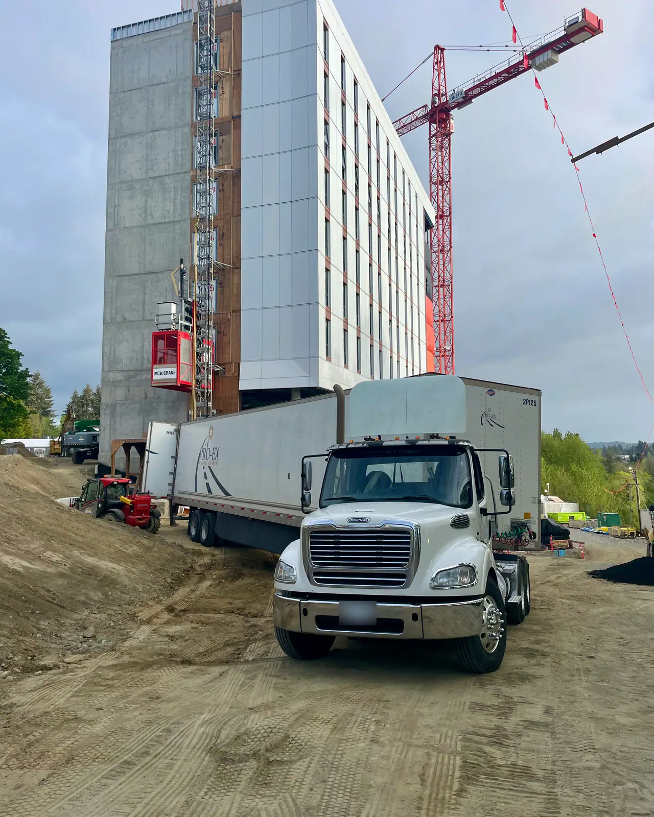 A white day-cab tractor coupled to a van trailer on the dirt apron of a tall building under construction, a red tower crane behind