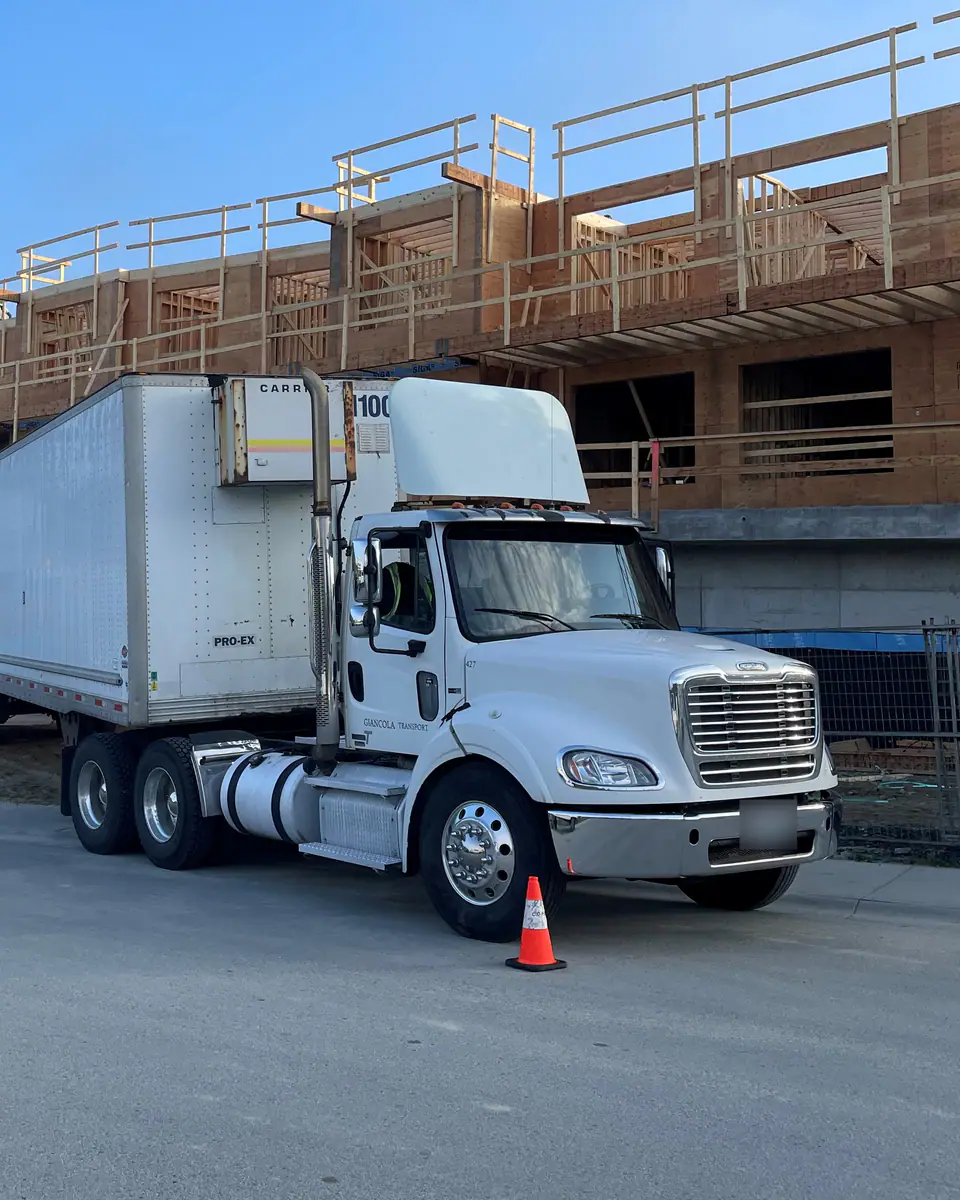 A white day-cab tractor and van trailer parked beside a timber-frame building under construction in early morning light