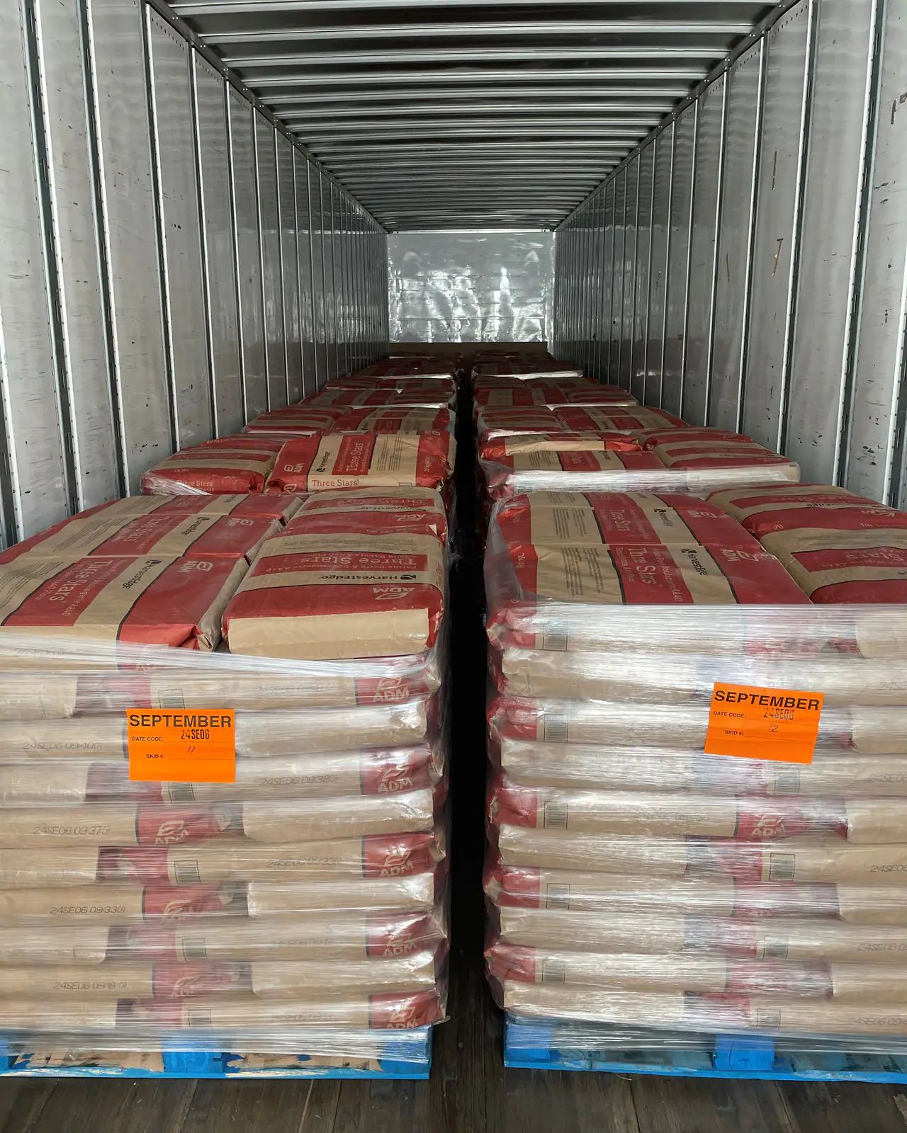 Shrink-wrapped pallets of bagged product with orange date labels, loaded two abreast down the length of a dry van trailer