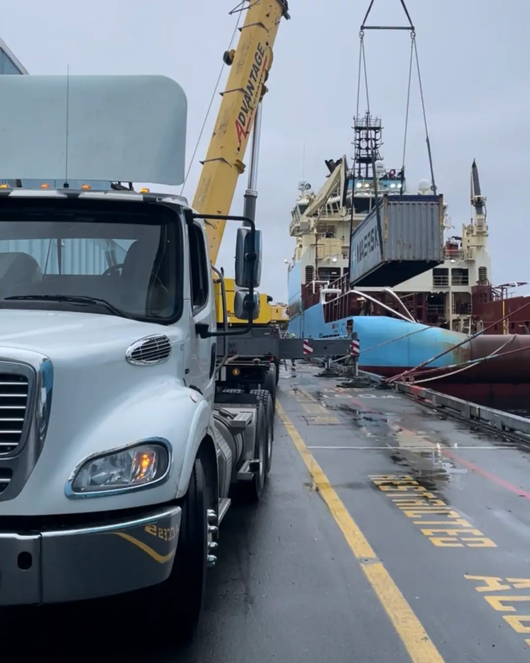 A dockside crane holds a shipping container on its hook beside a moored vessel, with a white tractor waiting on the wet wharf